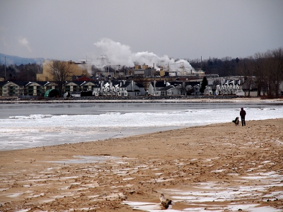 [Woman and dog walking the sandy beach beside the ice-laden waters. Billowing smoke stacks and houses are on the far shore.]